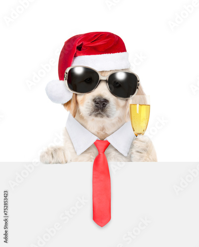 Young Golden retriever puppy wearing sunglasses, necktie and santa hat looks above empty white banner and holds glass of champagne. isolated on white background © Ermolaev Alexandr