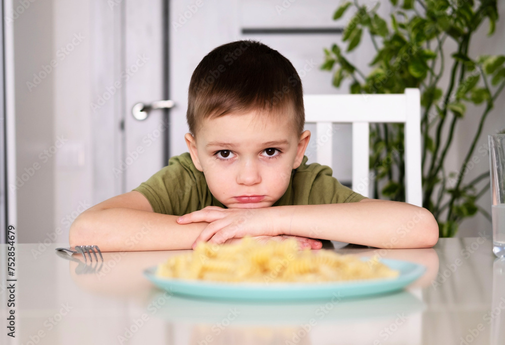 Poor little boy refuses to eat. An upset child sits in kitchen at table without an appetite and does not want to eat tasteless food. Avoiding pasta. Selective focus Poor little boy refuses to eat. An upset child sits in kitchen at table without an appetite and does not want to eat tasteless food. Avoiding pasta. Selective focus