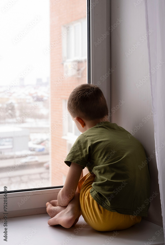 Sad cute child looks out window and feels sad. Boy is depressed. Vertical image Sad cute child looks out window and feels sad. Boy is depressed. Vertical image