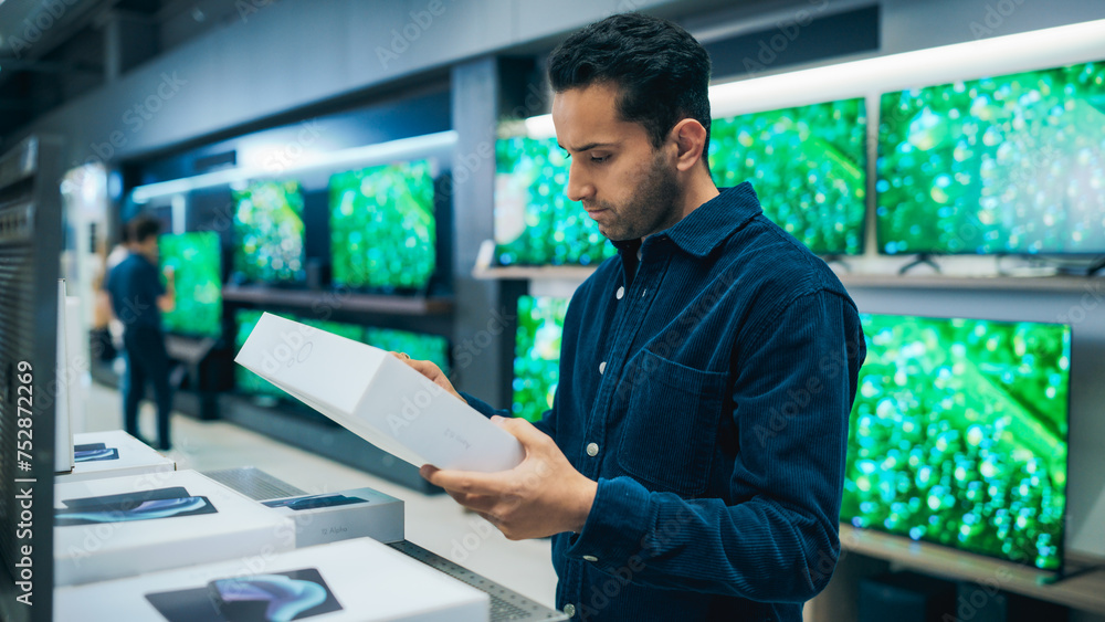 Handsome Young Man Shopping for a Tablet Computer in a Home Electronics Store. Multiethnic Customer Comparing Different Technology Features, Sale Price and Battery Life on a New Device Handsome Young Man Shopping for a Tablet Computer in a Home Electronics Store. Multiethnic Customer Comparing Different Technology Features, Sale Price and Battery Life on a New Device