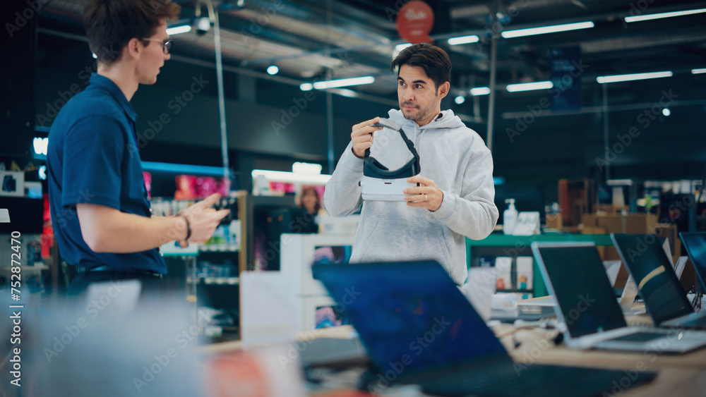 Young Man Having a Conversation with a Salesperson in a Electronics Store while Trying Out a Virtual Reality Device. Customer Wishes to Buy a VR Set. Shopper Explores Latest Options with Expert Advice Young Man Having a Conversation with a Salesperson in a Electronics Store while Trying Out a Virtual Reality Device. Customer Wishes to Buy a VR Set. Shopper Explores Latest Options with Expert Advice