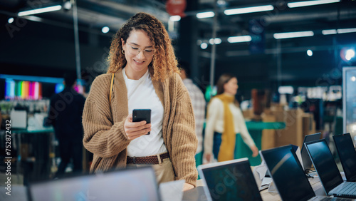 Portrait of a Happy Hispanic Woman Using a Smartphone, Texting with Friends on an Online Social Media App. Client Shopping for a Laptop Computer in a Home Electronics Store