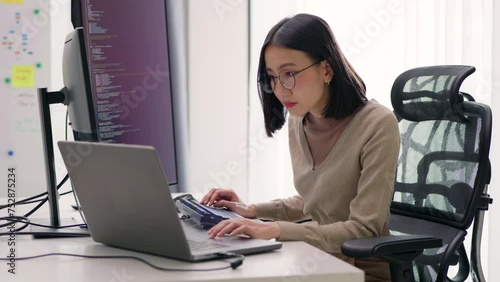 A focused female software developer programs at her workstation, her attention fixed on the large monitor. The office setting is modern and bright, indicative of a professional tech environment. photo