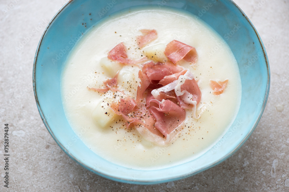 Middle closeup of potato soup served with prosciutto in a turquoise bowl, horizontal shot on a beige stone surface, selective focus Middle closeup of potato soup served with prosciutto in a turquoise bowl, horizontal shot on a beige stone surface, selective focus