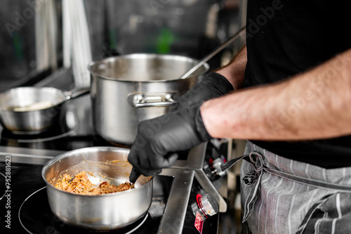 close-up of someone cooking meat in a stainless steel pan on a modern stove. The person wears black gloves for hygiene while handling food. The well-cooked meat has a brownish color. cooking process