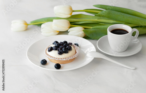 Panna cotta dessert with blueberries, cup of coffee, white tulip flowers bouquet on white marble table background, aesthetic sweet morning breakfast