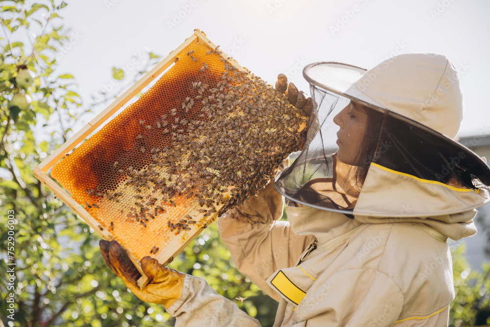 Happy smiling female Beekeeper in uniform standing in apiary and holding honeybee frame