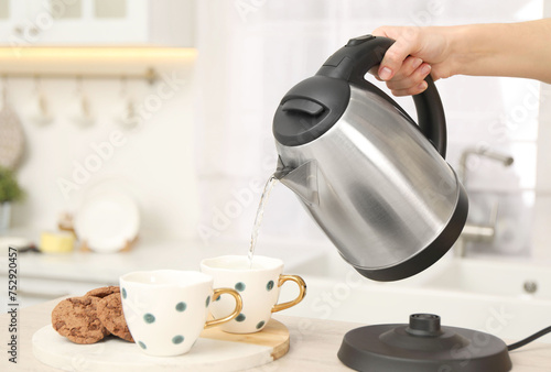 Woman pouring hot water from electric kettle into cup in kitchen, closeup. Space for text