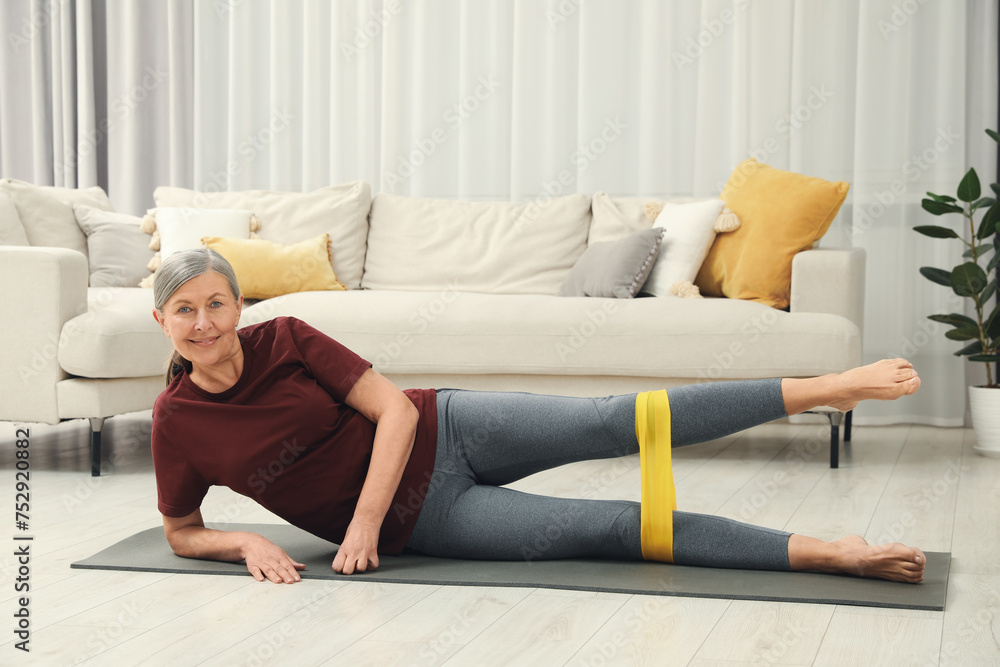 Fototapeta premium Senior woman doing exercise with fitness elastic band on mat at home