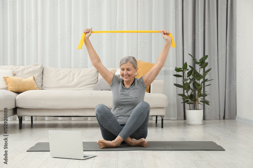 Senior woman doing exercise with fitness elastic band near laptop on mat at home
