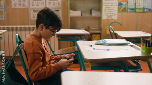 Child kid son pupil student bad behavior boy schoolboy playing video game on mobile phone at lesson at school hide smartphone under class desk distract from studying gadget addiction writing learning