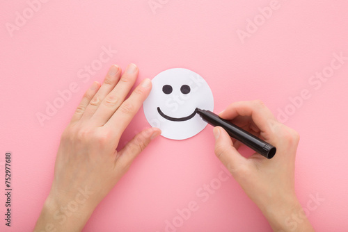 Young adult woman hand holding black marker and drawing happy smiling face on white paper on light pink table background. Pastel color. Closeup. Point of view shot. Top down view. photo