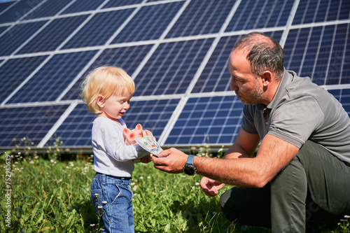 Father and his little son putting cash into piggy bank to save money on background of solar panels. Little kid interested in saving money for future. Concept of investment in green energy.
