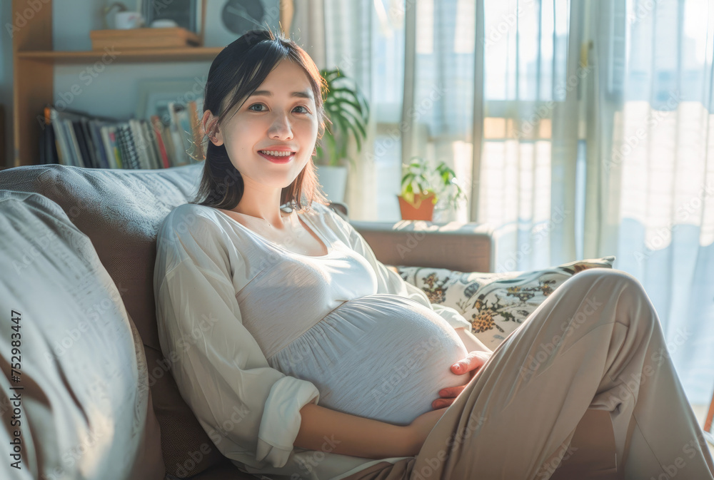 Calm young pregnant woman sitting on sofa and embracing her belly. Portrait of pregnant woman resting at her cozy home. Concept of motherhood and expecting baby. Calm young pregnant woman sitting on sofa and embracing her belly. Portrait of pregnant woman resting at her cozy home. Concept of motherhood and expecting baby.