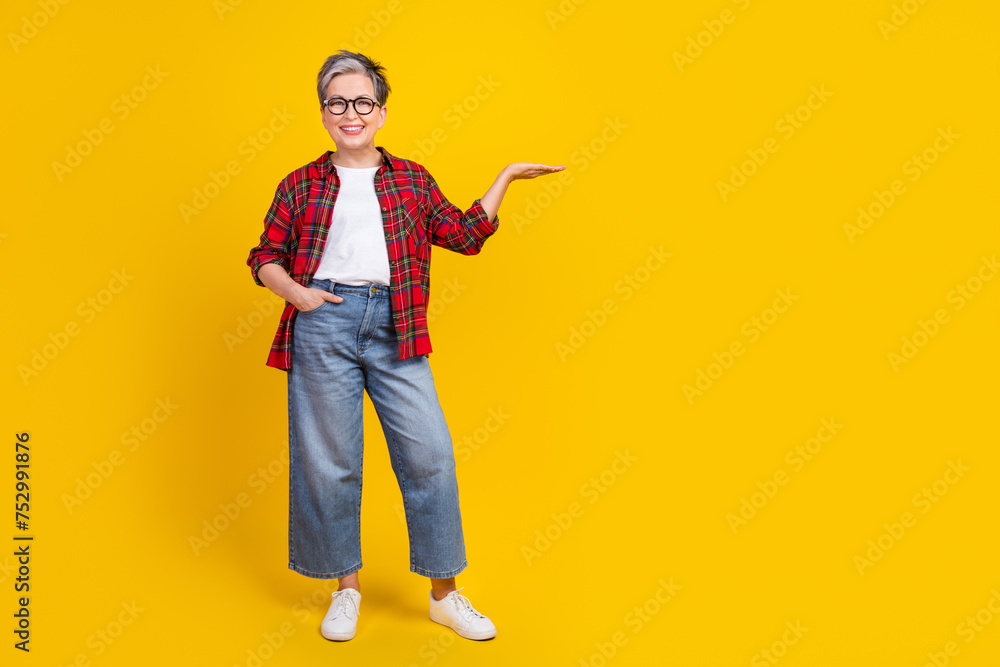 Full body photo of positive aged lady arm palm hold demonstrate empty space isolated on yellow color background Full body photo of positive aged lady arm palm hold demonstrate empty space isolated on yellow color background
