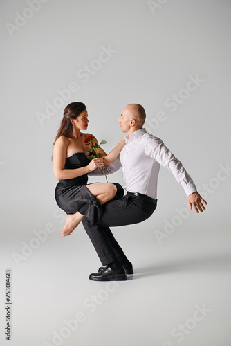 brunette woman in black dress holding red rose and balancing on laps of male dancer on grey backdrop