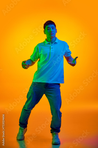 Confident young man in casual attire gesturing with open hands in vibrant neon light against gradient orange background. Concept of human emotions, fashion and beauty, self-expression, work and hobby.