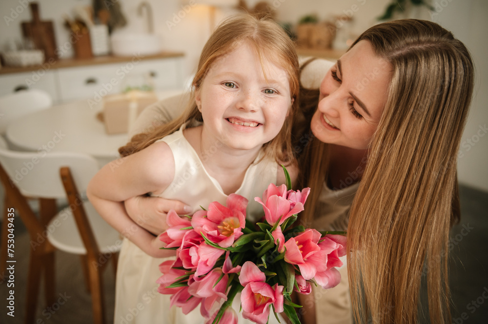 Mom and kid hugging, smiling. Happy family holiday. Cute little child girl greeting mother in kitchen. Beautiful daughter gives mother big bouquet of tulip flowers and makes surprise for Mother's Day. Mom and kid hugging, smiling. Happy family holiday. Cute little child girl greeting mother in kitchen. Beautiful daughter gives mother big bouquet of tulip flowers and makes surprise for Mother's Day.