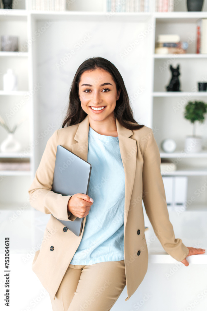 Vertical photo of successful arabian or indian young business woman in a suit, digital manager, seo, holding laptop while standing in a modern office space near work desk, looks and smile at camera Vertical photo of successful arabian or indian young business woman in a suit, digital manager, seo, holding laptop while standing in a modern office space near work desk, looks and smile at camera