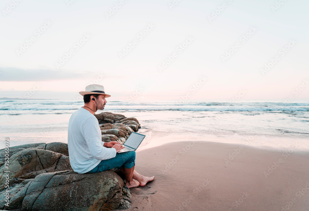 mature latin digital nomad sitting on the beach with a laptop working at sunset over the ocean	