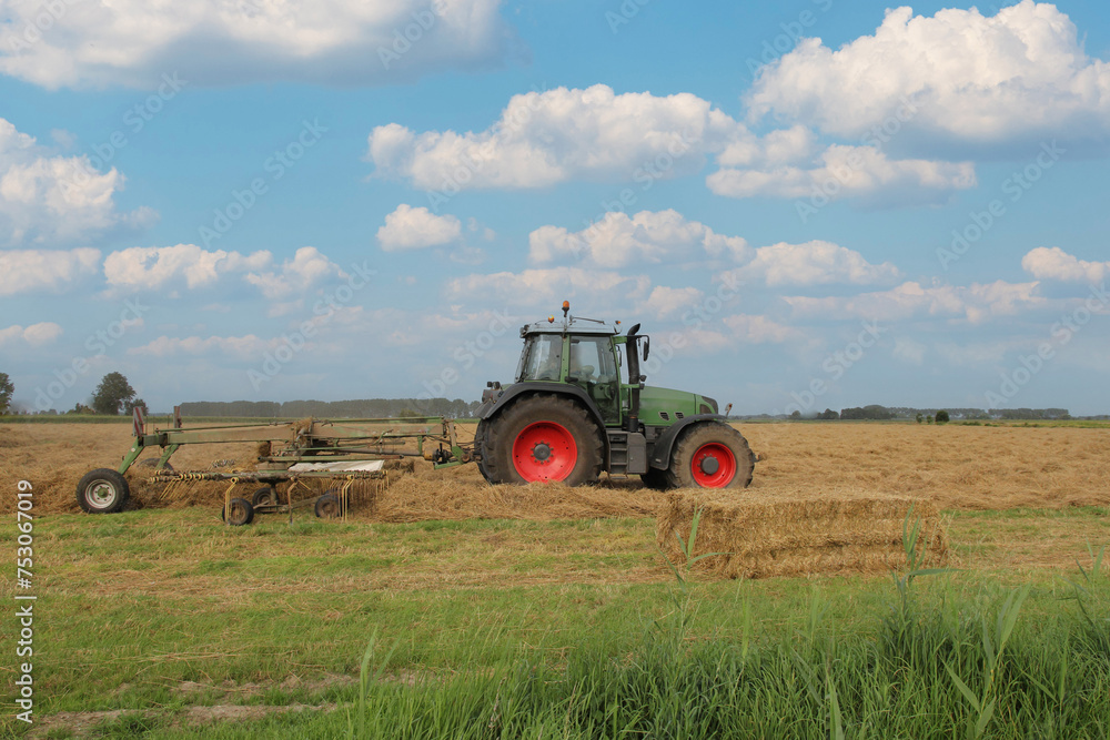 a farmer is driving with a tractor with a hay rake in a field with straw and a blue sky with clouds a farmer is driving with a tractor with a hay rake in a field with straw and a blue sky with clouds