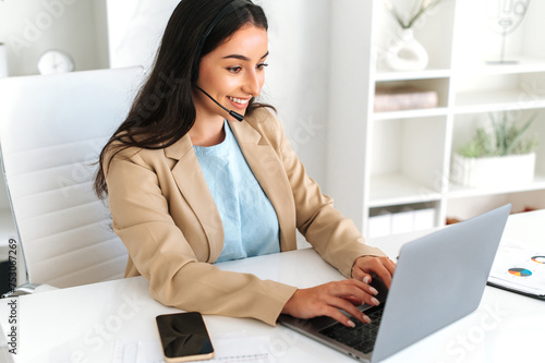 Call center, customer support service. Friendly positive woman, call center operator with headset, working on support hotline in a modern office with a laptop, looking at the screen, smiling