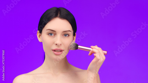 Close-up portrait of an attractive beautiful young topless woman standing isolated on a purple background with a makeup brush. Concept of cosmetology, cosmetic care products. photo