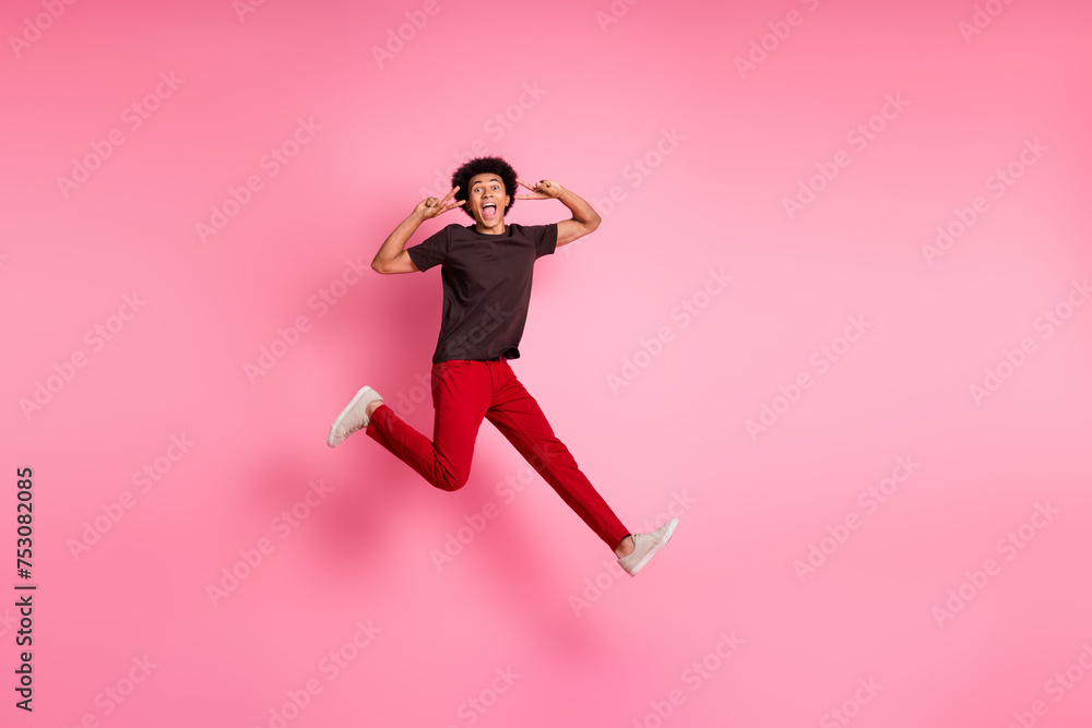 Full body length photo of young funny curly hair sportive guy jumping trampoline showing double v sign isolated on pink color background