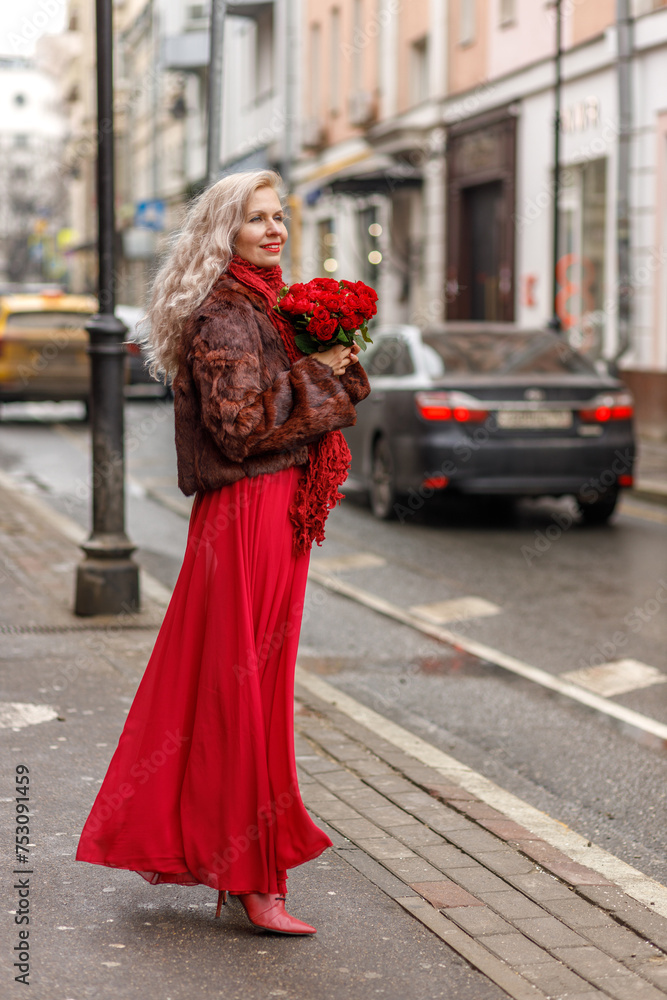 Obraz premium An elegant blonde in a red evening dress and fur coat is standing on the streets of the city, waiting for a taxi. Spring concept, international women's day. March 8