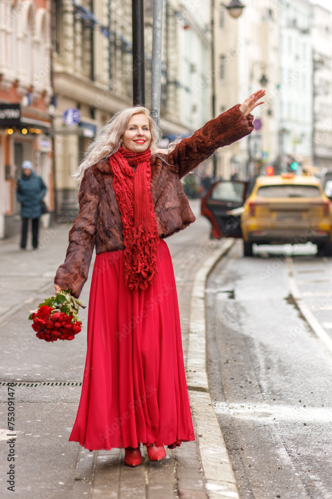 Obraz premium An elegant blonde in a red evening dress and fur coat is standing on the streets of the city, waiting for a taxi. Spring concept, international women's day. March 8