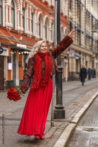 An elegant blonde in a red evening dress and fur coat is standing on the streets of the city, waiting for a taxi. Spring concept, international women's day. March 8 © Светлана Густова