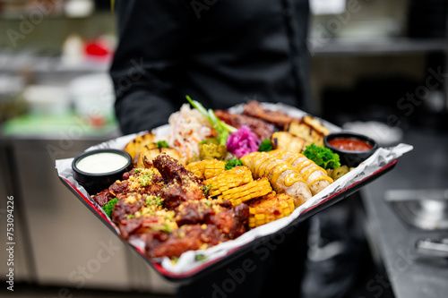 A server in black attire holds a tray of assorted delicious food, including grilled meat, corn, and sauces, in a professional kitchen setting