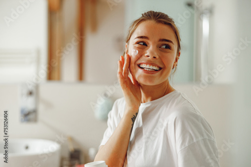 Murais de parede Happy woman applying hydrating moisturizer on her face sitting in bathroom
