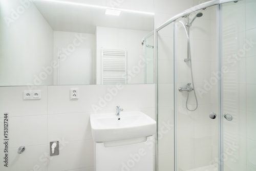 Bright white small bathroom with sink and shower. The bathroom has a large mirror with led tape, several electrical sockets, heating ladder and water with waste for the washing machine.