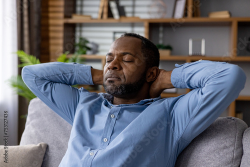 Serene African American man with closed eyes, enjoying tranquility while sitting on a sofa in a cozy living room setting.