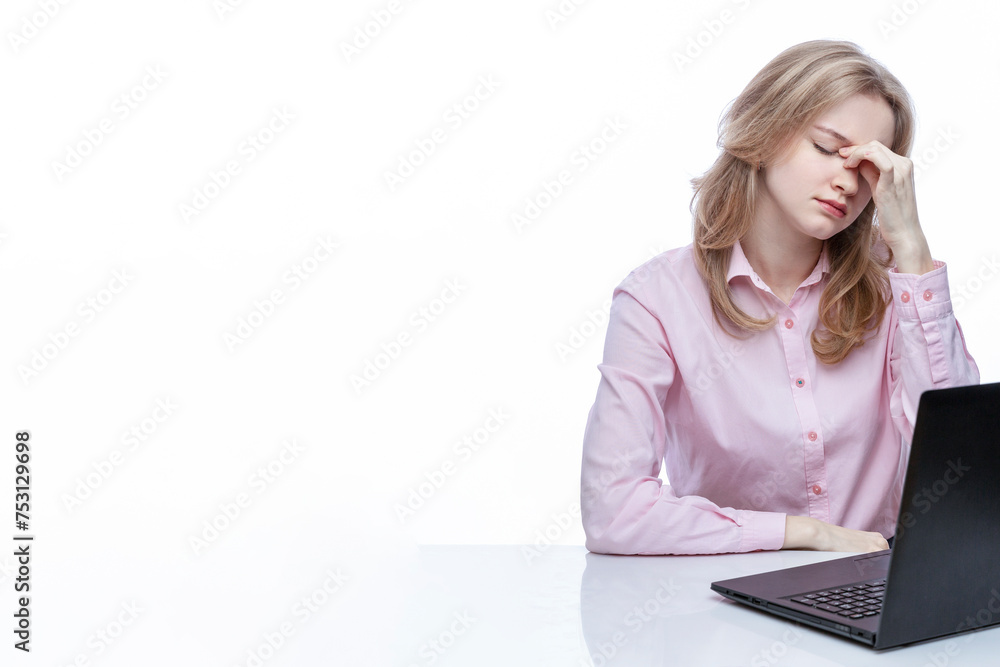 A young tired girl sits at a table in front of a laptop and touches her eyes with her fingers. Cute blonde girl in a pink shirt Work, study and stress. Isolated on a white background. Space for text. A young tired girl sits at a table in front of a laptop and touches her eyes with her fingers. Cute blonde girl in a pink shirt Work, study and stress. Isolated on a white background. Space for text.