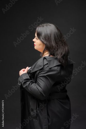 A woman in a black coat stands in front of a black background. She is wearing a red lipstick and has a serious expression on her face