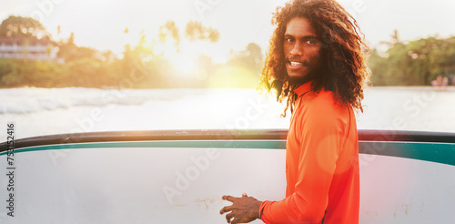 Portrait of black long-haired teen boy with surfboard ready for surfing with sunset backlight. He walking in Indian ocean waves. Extreme water sports and exotic countries concept, Udawalawe, Sri Lanka © Soloviova Liudmyla