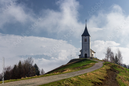 A path leading toward the catholic church. An ancient church on the clean grassy hill with fog surrounding it. Church of saint Primoz and Felician - Jamnik - Slovenia. 