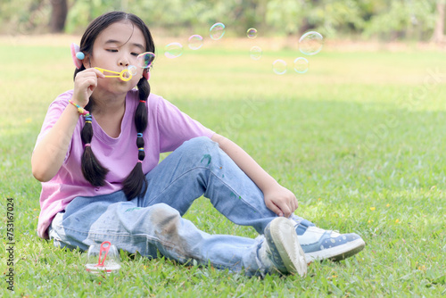 Happy cute Asian girl with pigtails blowing soap bubbles while sitting on green grass in nature garden park. Kid spending time outdoors in meadow. Cute child having fun with bubble in summer park. #753167024