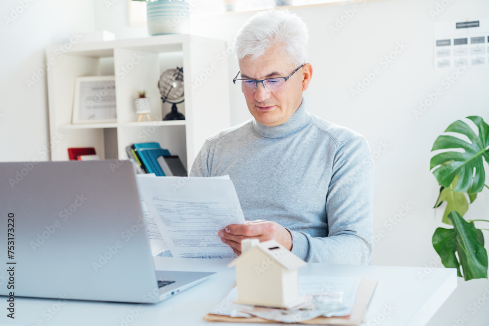 Middle aged man working on laptop with house model money box and docs on table. Housing cost and payments. Real estate, property investment. Home mortgage loan rate. Saving money for retirement. Middle aged man working on laptop with house model money box and docs on table. Housing cost and payments. Real estate, property investment. Home mortgage loan rate. Saving money for retirement.