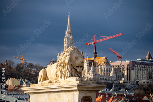 Lion statue on famous Chain Bridge in the city of Budapest, Hungary with Matthias Church and Fisherman`s Bastion rising in the distance