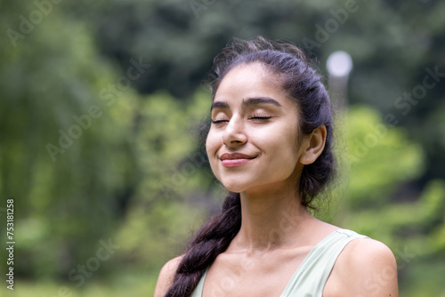 Close-up photo of a smiling and relaxed young Indian woman standing outside in a park and resting after a run, eyes closed resting and meditating