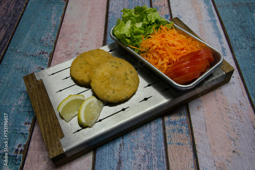 Top view of two fried fish burgers with lemon and a lettuce, carrot and tomato salad, served in a metal board on the rustic wooden table.	