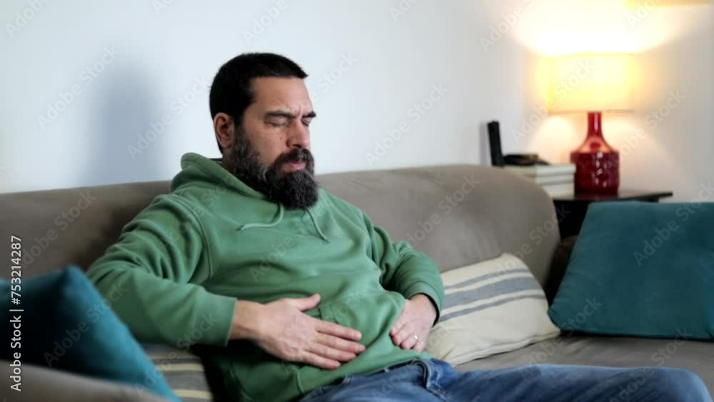 Bearded man grimacing in pain, suffering from stomach ache sitting on sofa at home clutching his stomach due to severe pain. Bearded man grimacing in pain, suffering from stomach ache sitting on sofa at home clutching his stomach due to severe pain.