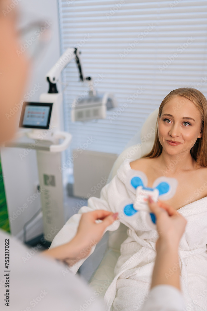 Vertical shot of unrecognizable female cardiologist attaching holter sensors on smiling woman patient with heart disease. Young woman during functional rehabilitation in cardiology room in clinic. Vertical shot of unrecognizable female cardiologist attaching holter sensors on smiling woman patient with heart disease. Young woman during functional rehabilitation in cardiology room in clinic.