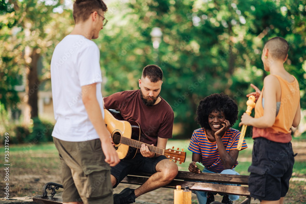 Four multiracial young people sitting in a park and playing guitar.