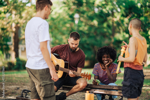Four multiracial young people sitting in a park and playing guitar.