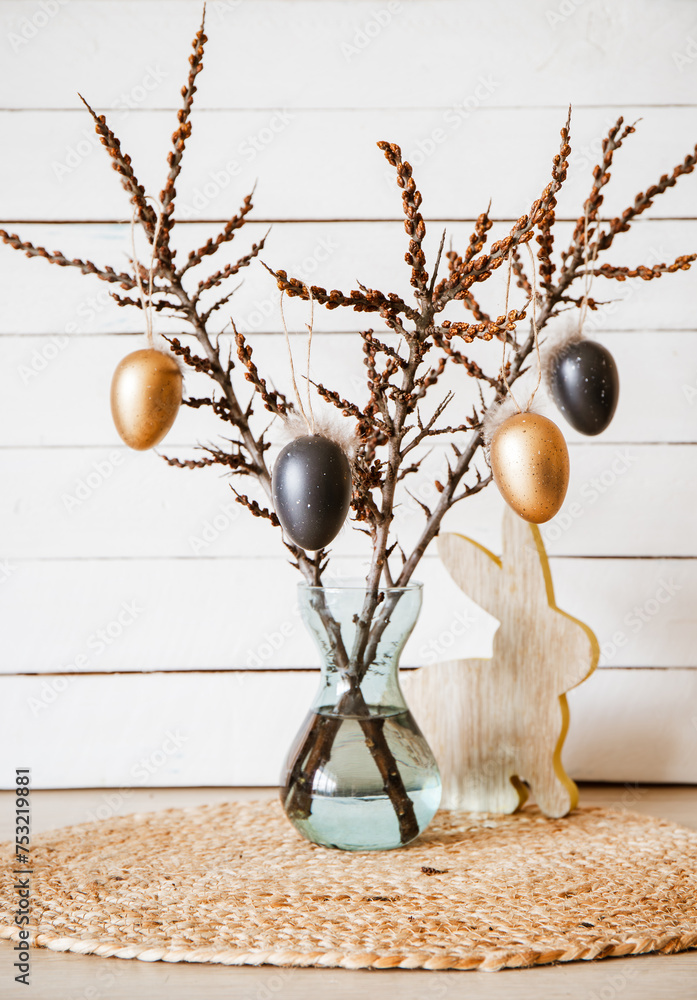 Tree branches in vase with black and bronze colored Easter eggs hanging on string, white rustic wood board background. Minimal home Easter decoration arrangement.