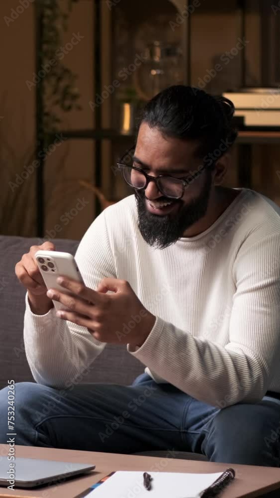Positive Indian man engages with phone, sharing laughter while seated on the sofa in the living room. Joyful expressions create a cheerful atmosphere in the tranquil setting of the night. Positive Indian man engages with phone, sharing laughter while seated on the sofa in the living room. Joyful expressions create a cheerful atmosphere in the tranquil setting of the night.